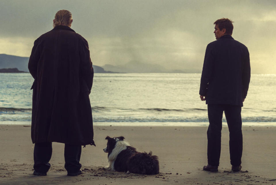 A still from the movie "The Banshees of Inisherin." Two men stand on a beach looking out into the water. A dog lays next to the older man.