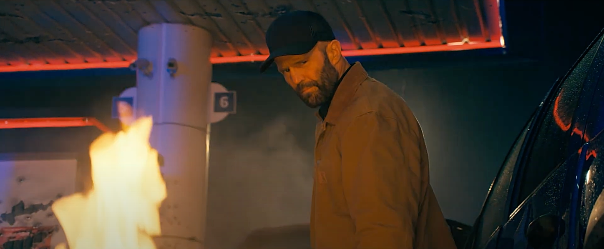 jason statham in the beekeeper, wearing a black baseball cap and dark khaki jacket, staring at a fire next to a vehicle