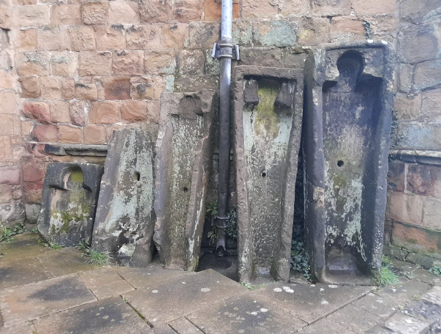 Five ancient stone stocks of varying sizes against a brick wall, possibly historic punishment devices