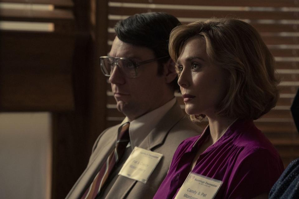 a publicity photo from the miniseries love and death, featuring actors patrick fugit and elizabeth olsen sitting in a room, looking straight ahead, with fugit wearing a tan suit and glasses, and olsen wearing a purple shirt