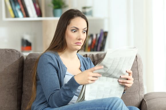 Young woman reading a newspaper with wide eyes and a furrowed brow.