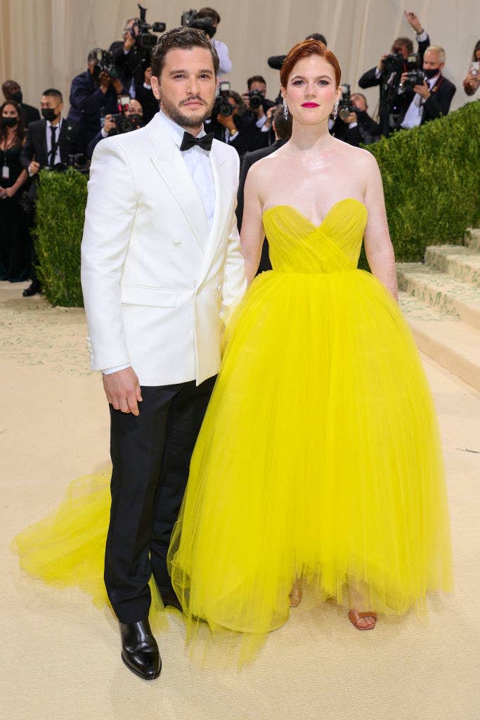 Kit Harrington wears a light colored blazer over dark slacks and Rose Leslie wears a strapless floor length brightly colored gown