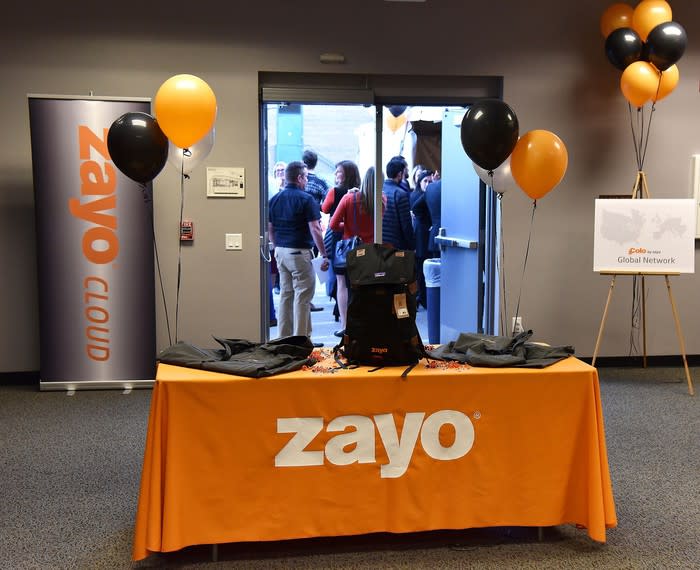 Desk with Zayo logo and orange and black balloons, with a huddle of people in background.