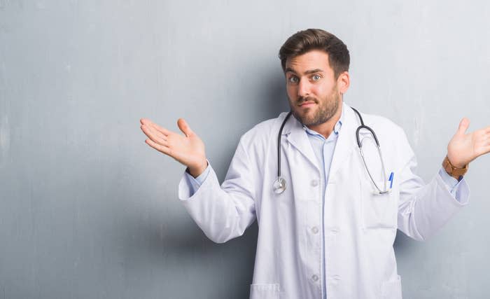 A man wearing a doctor's white lab coat and stethoscope shrugs with a confused expression