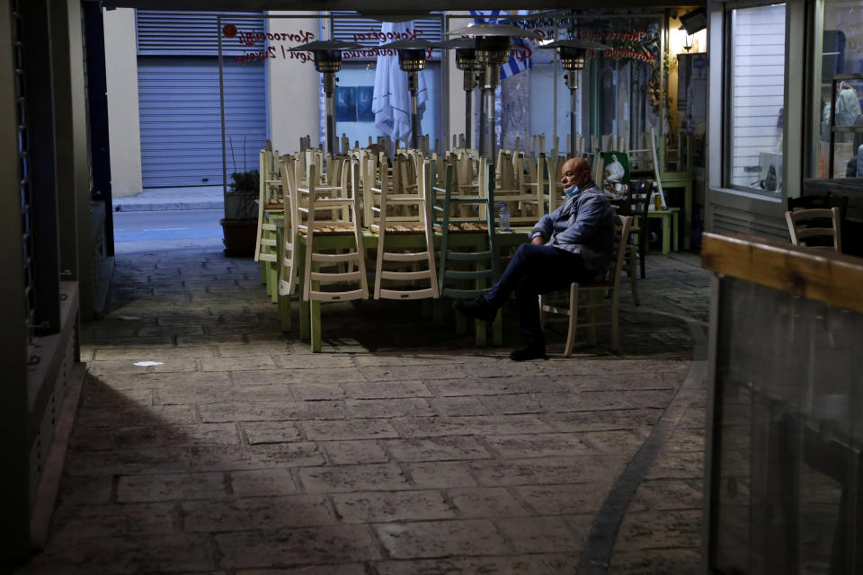 A man wearing a face mask sits by stacked chairs outside of a closed restaurant in central Nicosia, Cyprus, Thursday, Feb. 25, 2021. Cyprus will send all high school students back to their classrooms and open up gyms, pools, dance academies and art galleries next week in a further, incremental easing of the country's second nationwide COVID-19 lockdown. (AP Photo/Petros Karadjias)