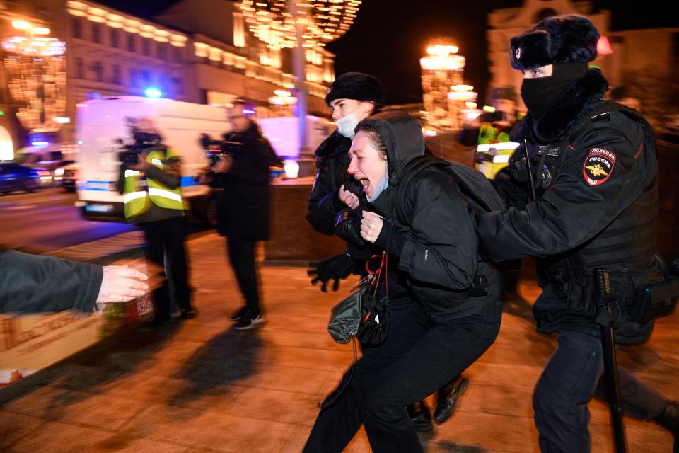 In a brightly lit street, a Russian police officer wearing a face covering with only his eyes visible grabs from behind a women demonstrator who is screaming in anguish.