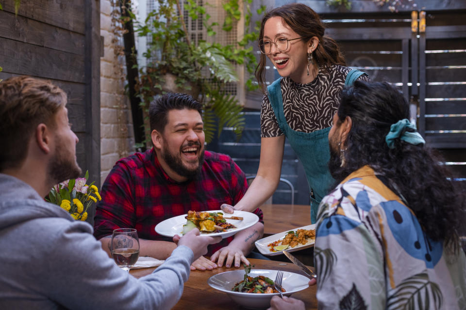 Four friends enjoying a meal together, one woman serving food, all smiling and engaged in conversation