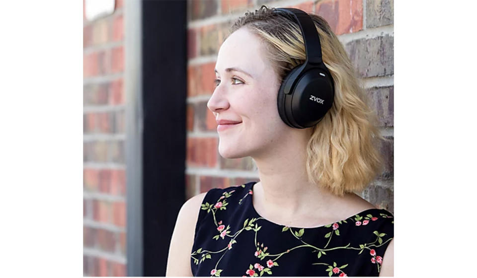 Smiling blonde woman wearing black ZVOX headphones leaning against brick wall