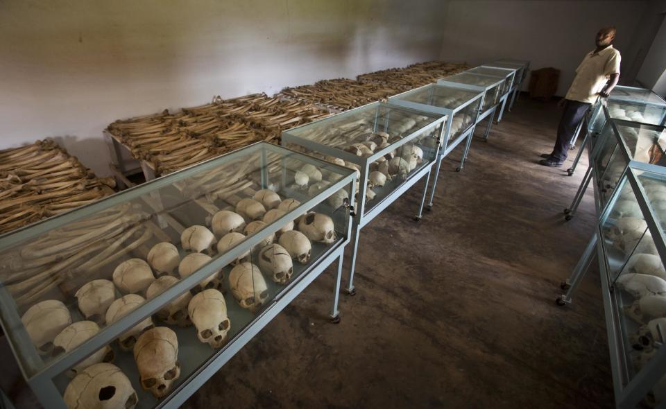 In this photo taken Thursday, March 27, 2014, Mike Nkuzumuwami stands by the rows of human skulls and bones that form a memorial to those who died in the redbrick church that was the scene of a massacre during the 1994 genocide, and which he helps to look after, in the village of Nyarubuye, eastern Rwanda. Rwandans gathered in the nearby town of Kirehe Thursday to watch the arrival of a small flame, symbolic fire traveling the country as Rwanda prepares to mark 20 years since ethnic Hutu extremists killed neighbors, friends and family during a three-month rampage of violence aimed at ethnic Tutsis and some moderate Hutus, the death toll of which Rwanda puts at 1,000,050. (AP Photo/Ben Curtis)