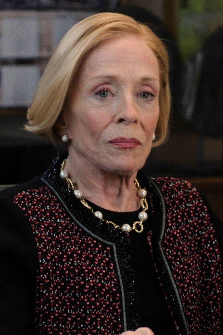 Holland Taylor sits at a conference table, dressed in formal attire, in a scene from a TV show