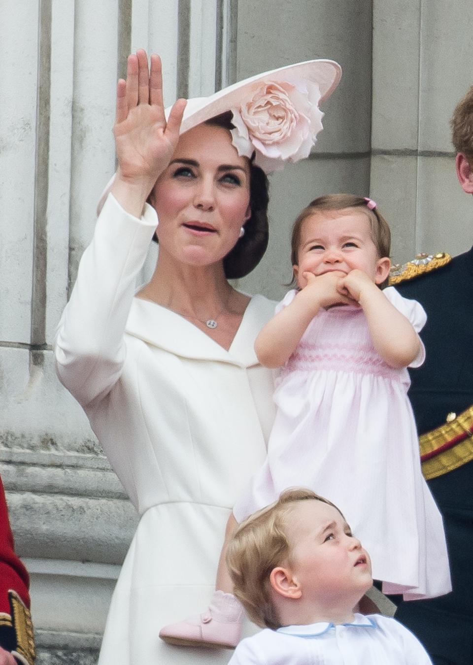 Kate Middleton holds Princess Charlotte and waves in front of a stone wall. Prince George looks up at the sky in the bottom of the frame.