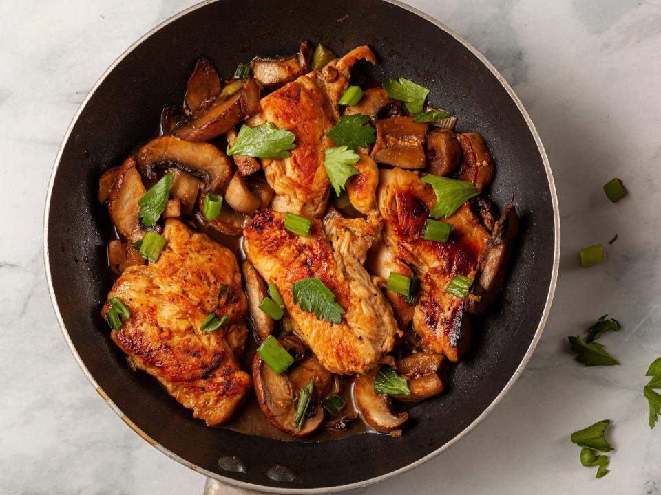 A nonstick frying pan with a chicken breast sautee inside. Meat is seasoned with herbs, tomato, spices, parsley, spring onions and mushrooms.