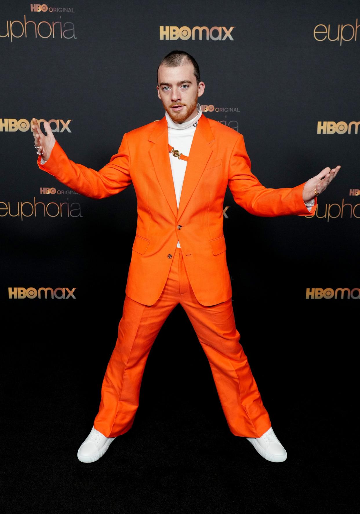 Angus in a bright orange suit with a white turtle neck, orange harness across his chest, and white sneakers.