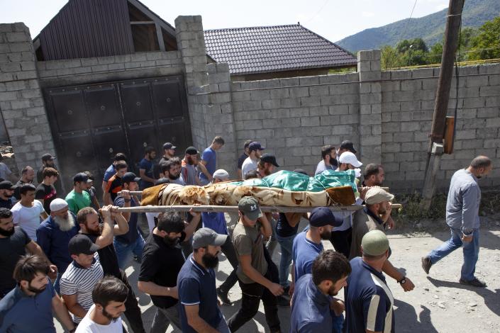 A crowd of men, one wearing a long beard and a skull cap, the rest young men in baseball caps, surround pallbearers carrying on their shoulders a dead body wrapped in a multicolored blanket and bound with rope to a stretcher.