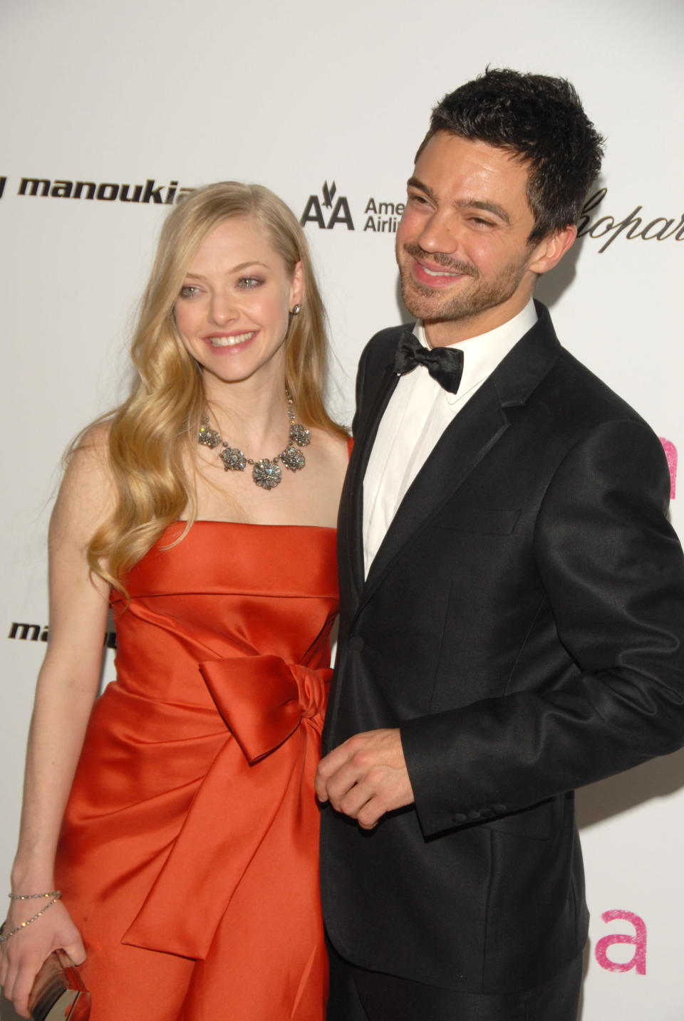 Amanda Seyfried and Dominic Cooper with their arms around one another smiling at the crowd at a red carpet event