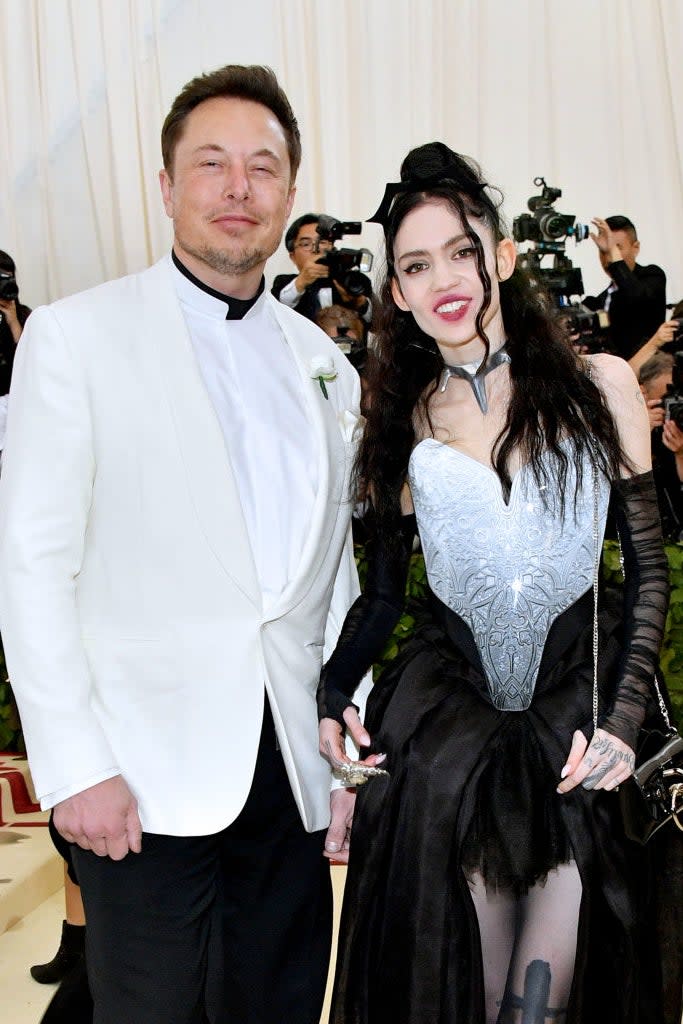 Elon Musk in a white suit stands with Grimes in a black and silver gown, both posing for the camera