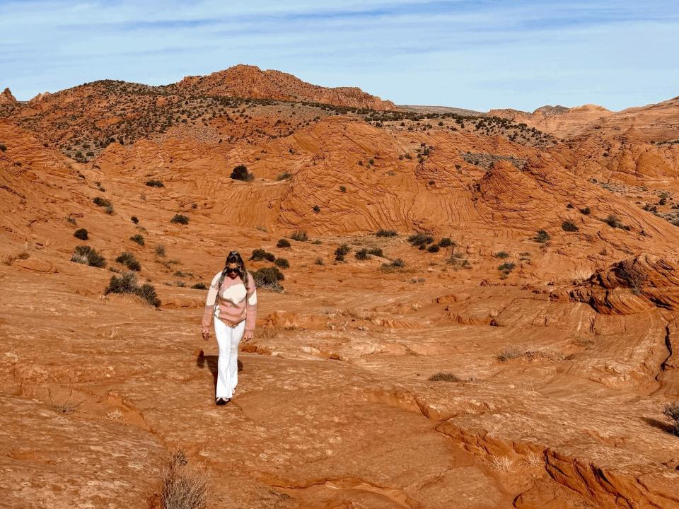 The writer wears white pants and a pink and white sweater and walks among red rocks and bushes with blue sky overhead