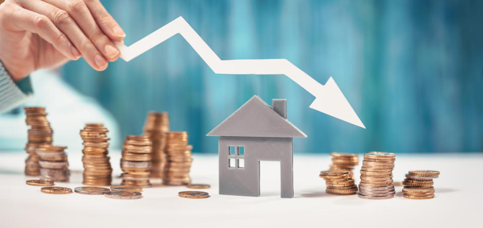 A hand holds a downward trending arrow above a small house model and stacks of coins, symbolizing a decline in the real estate market or home values