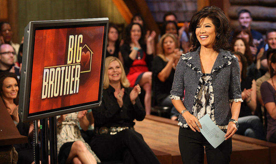 Julie Chen stands next to 'Big Brother' sign, hosting a show, audience in background