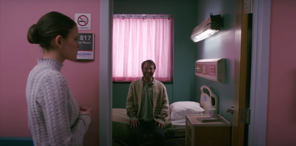 A woman passes a hospital room with a man inside smiling