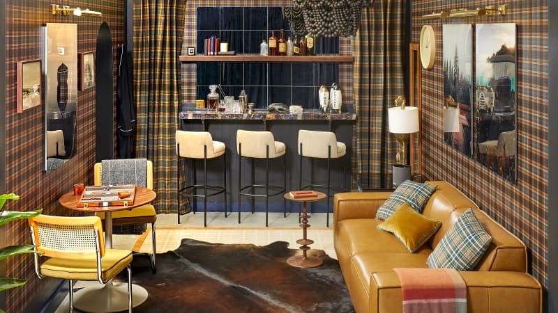 Head on view of a living room with black and yellow plaid wallpaper, a light brown leather couch and a wall of mirrors with a floating bar top and shelves against the wall.