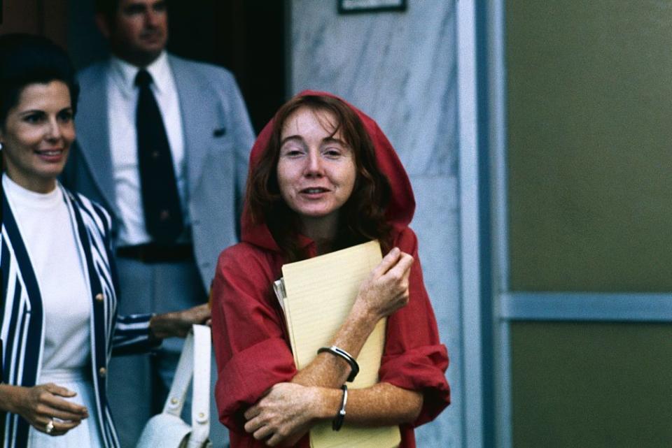 a woman in a red hooded jumpsuit being led in handcuffs out of a courthouse, holding a pad and papers