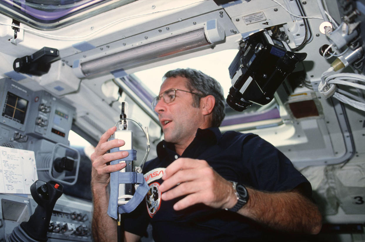  A man in a dark shirt holds what looks like a cordless phone inside the cockpit of a spacecraft. 