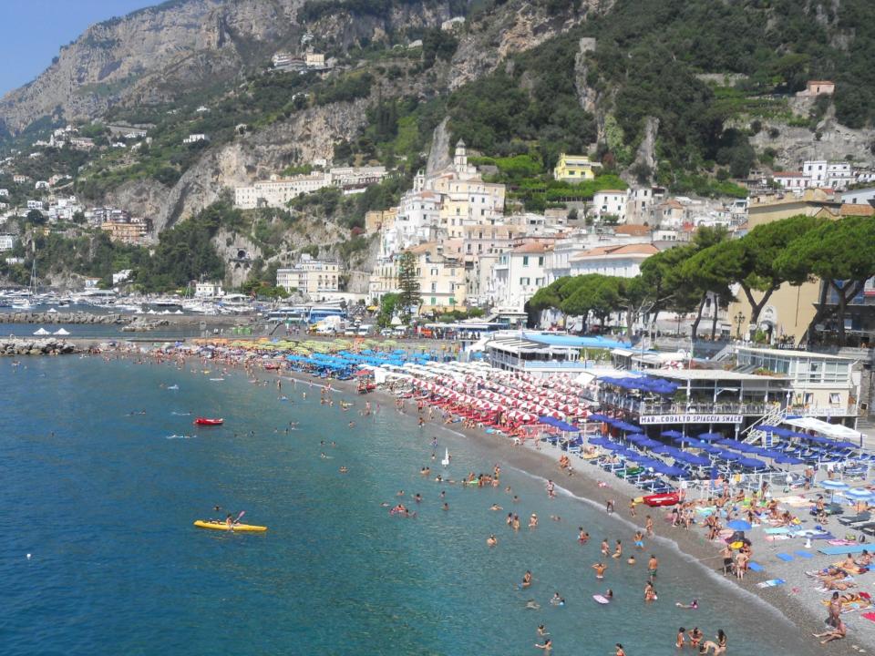 People swimming in the sea, and beach chairs and umbrellas lined up on the shore. Behind the beach are buildings and mountains.