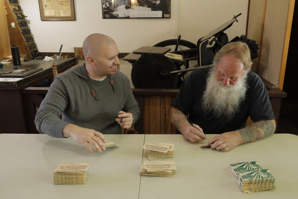 In this May 21, 2020 photo, printer Loren Ackerman, right, and Wayne Fournier, left, mayor of Tenino, Wash., sign pieces of wooden money Ackerman printed on an 1890s-era press in Tenino, Wash. In an effort to help residents and local merchants alike get through the economic fallout of the coronavirus pandemic, the small town has issued wooden currency for residents to spend at local businesses, decades after it created a similar program during the Great Depression. (AP Photo/Ted S. Warren)