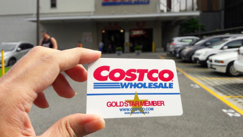 A person holds a Costco membership card in front of a Costco store.