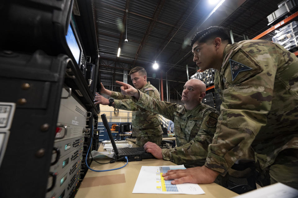 three military personnel in camouflage uniforms point at a computer screen in a hangar