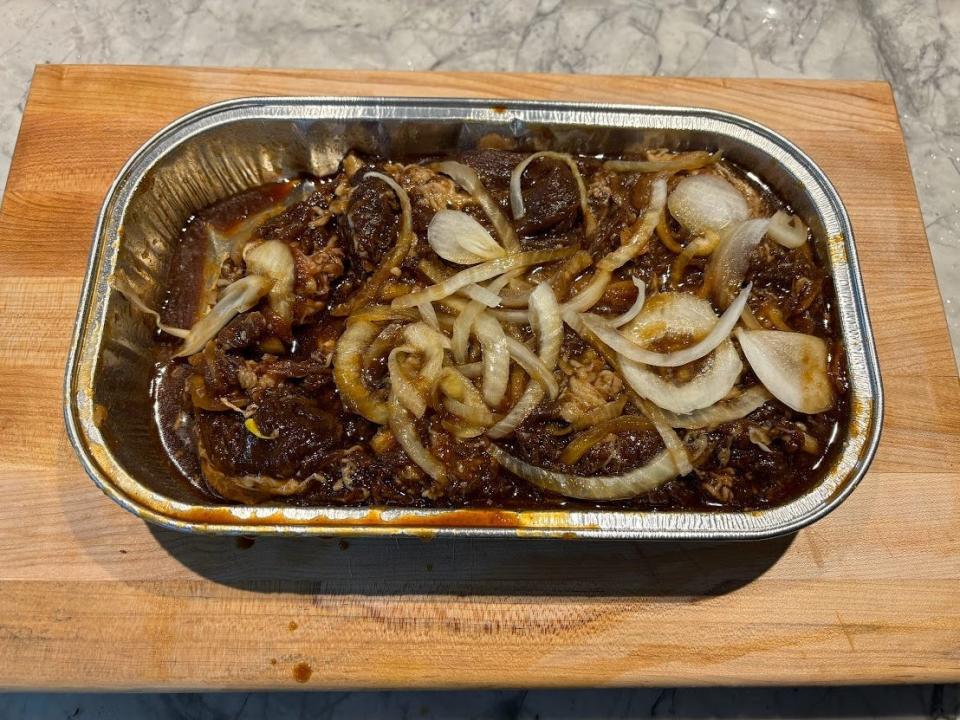 Raw beef bulgogi covered in a brown sauce with pieces of brown sauce in a tin tray on a wooden cutting board