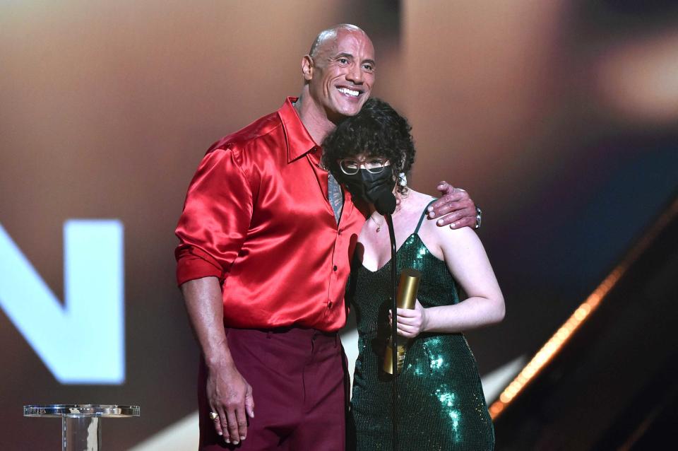 Johnson in a red silky shirt puts his left arm around a teen in a green sparkly dress as she holds his award in her left hand. (Getty Images)