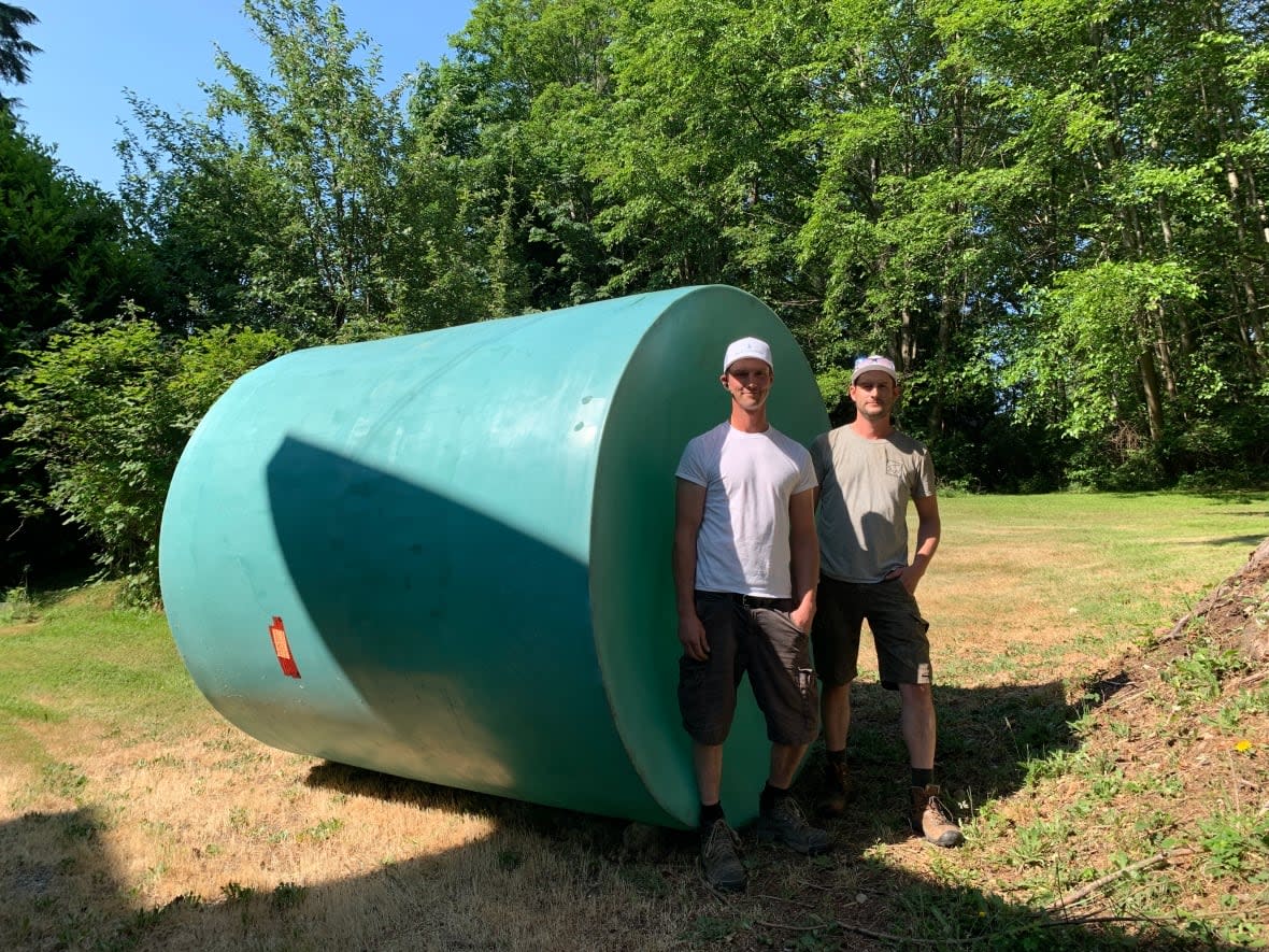 Gord, left, and Geoff Sloan stand in front of a 9,000-litre tank they are installing at a property in Sechelt, B.C., to collect rainwater for irrigation and non-drinking uses. The brothers' Sunshine Coast business is booming as more hot, dry weather arrives in a region dogged by drought for the past few years. (Yvette Brend/CBC - image credit)