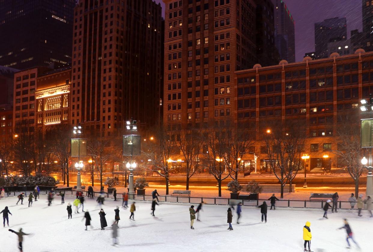 People ice skating on an ice rink during late evening in Chicago, with a row of buildings behind, during Christmas season, lights cast an orange glow but rink is brightly lit