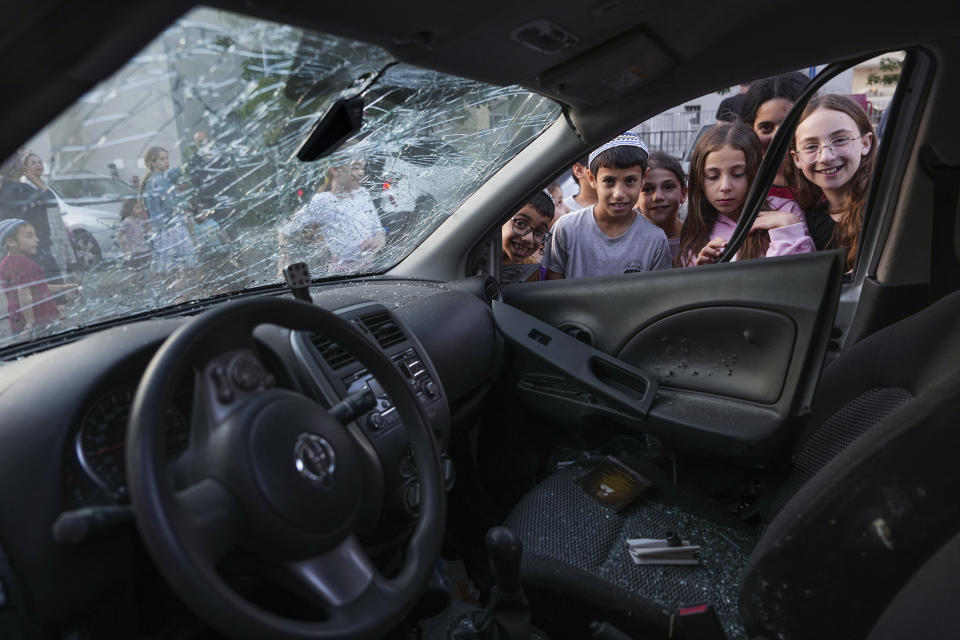 Israeli kids smile at the camera as others inspect a car struck by rocket fire from the Gaza Strip, in the southern Israeli city of Sderot, Tuesday, May 2, 2023. The Israeli military says that Palestinian militants in Gaza have fired a barrage of rockets following the death of Khader Adnan, a high-profile Palestinian prisoner in Israeli custody after a nearly three-month-long hunger strike. (AP Photo/Tsafrir Abayov)