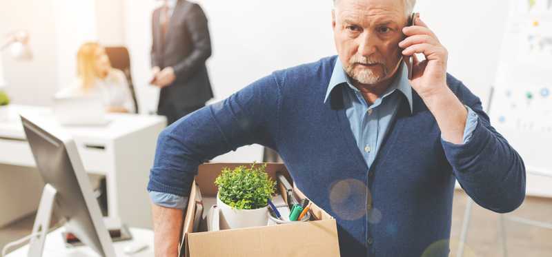 Older man on a cell phone leaving an office with personal belongings in a box.