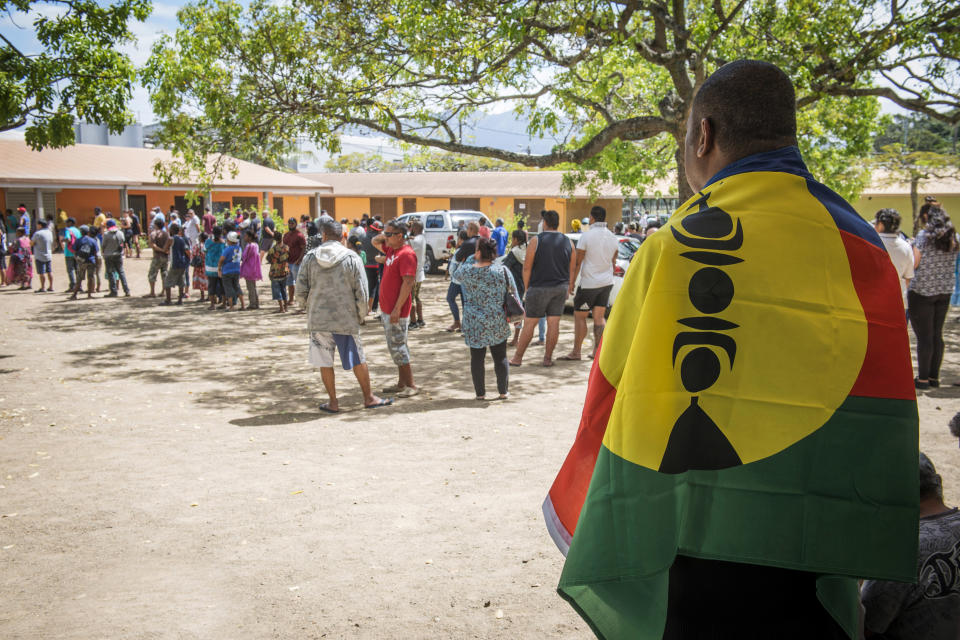 A man drapes his country's flag over his shoulders as residents of New Caledonia's capital, Noumea, wait in line at a polling station before casting their vote as part of an independence referendum, Sunday, Nov. 4, 2018. Voters in New Caledonia are deciding whether the French territory in the South Pacific should break free from the European country that claimed it in the mid-19th century. (AP Photo/Mathurin Derel)