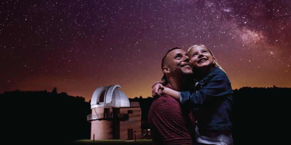 A father and daughter gaze up at the stars in front of the Monteville Observatory Park in Ohio. The sky is purple and orange and filled with brilliant stars.