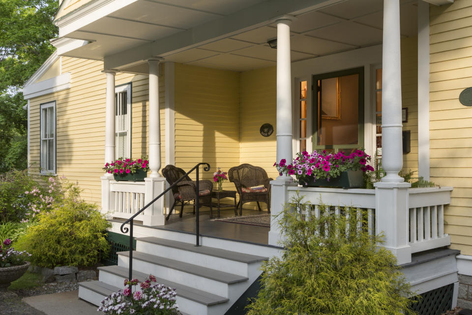 A cozy front porch with wicker chairs, flower boxes, and blooming plants. A welcoming space at the entrance of a yellow house