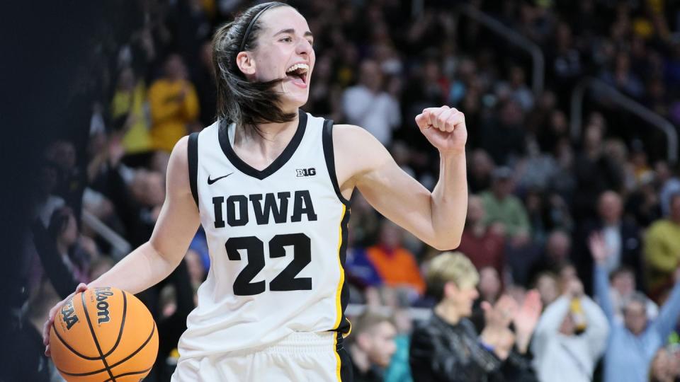 caitlin clark holds a basketball in one hand and pumps her other fist, she wears a white and black basketball uniform for iowa and smiles