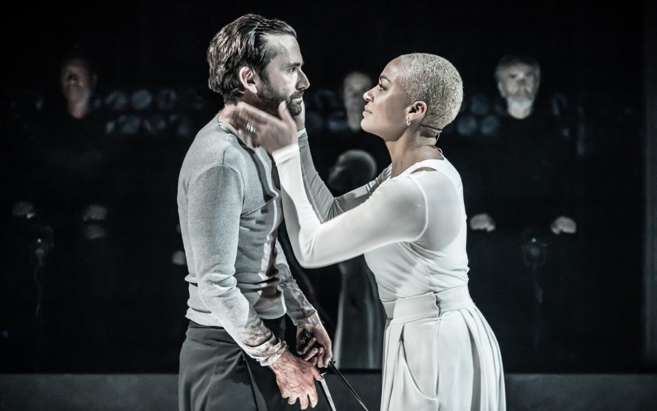 David Tennant and Cush Jumbo on stage in sombre clothes with figures seen in the darkness behind them