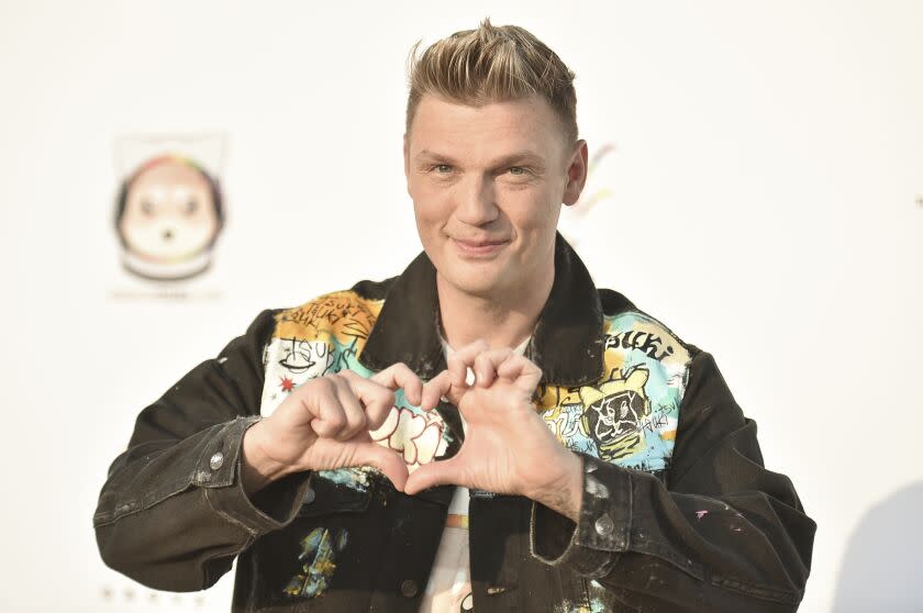 A man with short blond hair wearing a jean jacket and forming a heart shape with his hands