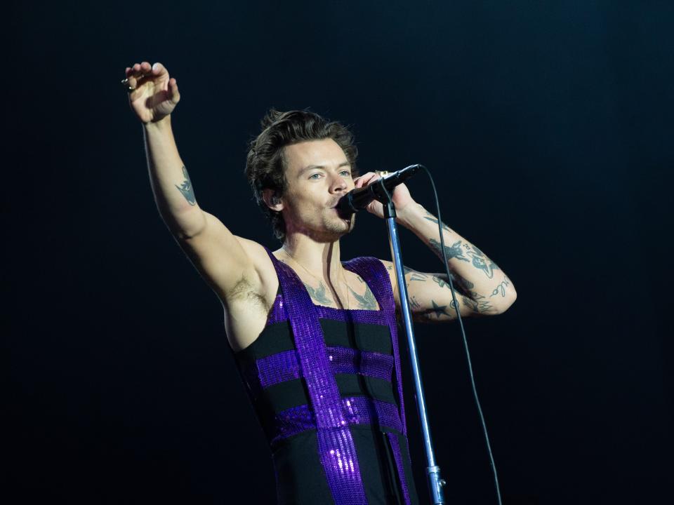 Harry Styles on stage holding a microphone in a purple jumpsuit