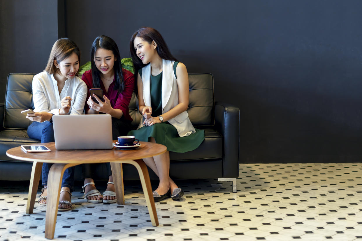 Group of charming beautiful Asian women using smartphone and laptop, chatting on sofa at cafe, modern lifestyle with gadget technology or working woman on casual business concept