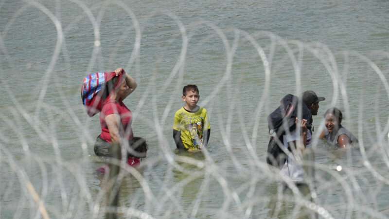 Migrants wading through water near a razor wire barrier