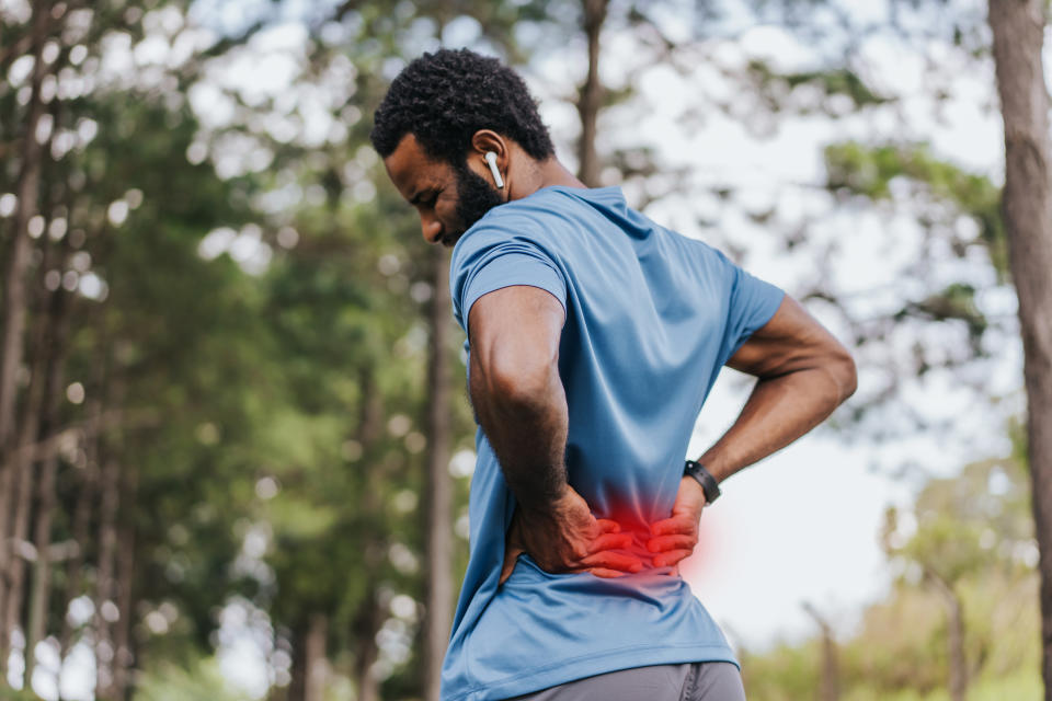 Person holding lower back in discomfort, wearing athletic clothing and earbuds, standing outdoors among trees