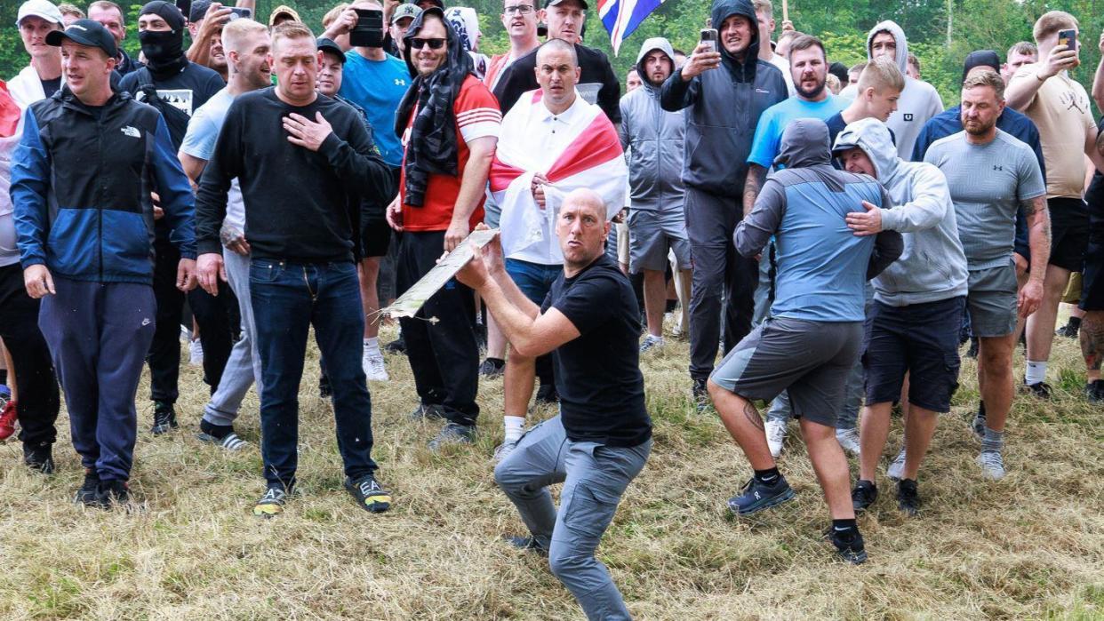 A group of men are gathered some holding mobile phones, some have masks on. At the forefront is man in a black t-shirt and jeans holding a thin piece of wood in a way which looks like he is about to throw it.