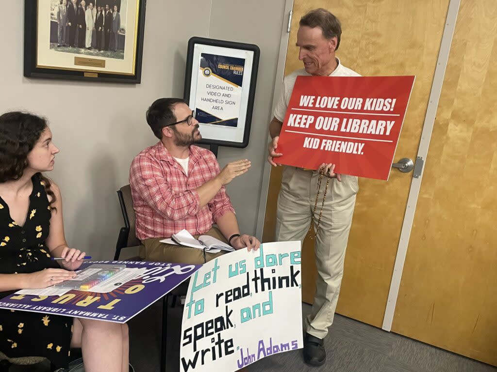 Opponents and a supporter of restrictions on library content, each holding signs backing their stance, talk before a St. Tammany Parish Council meeting