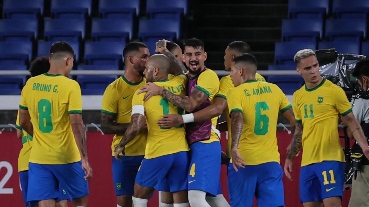 Brazil's players are huddled together celebrating a goal. They are wearing yellow shirts with green lettering and blue shorts 